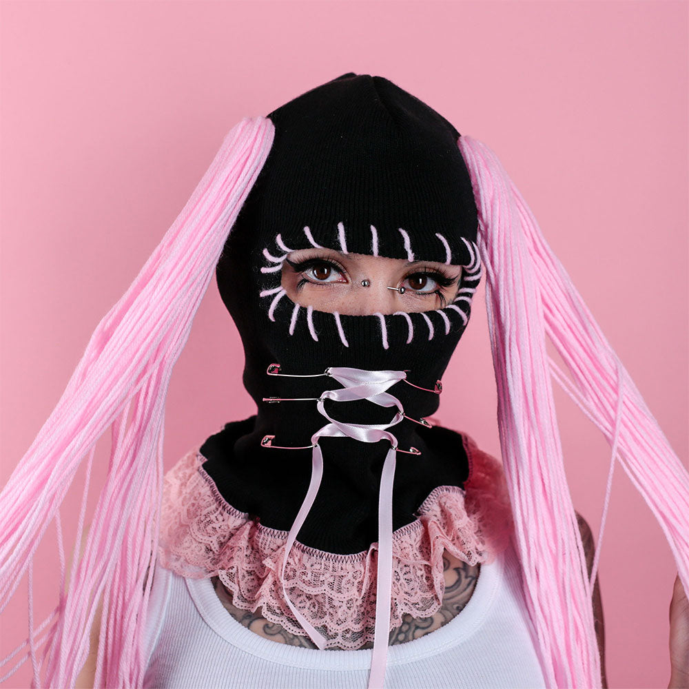 Person wearing a black balaclava with pink hair extensions against a pink background