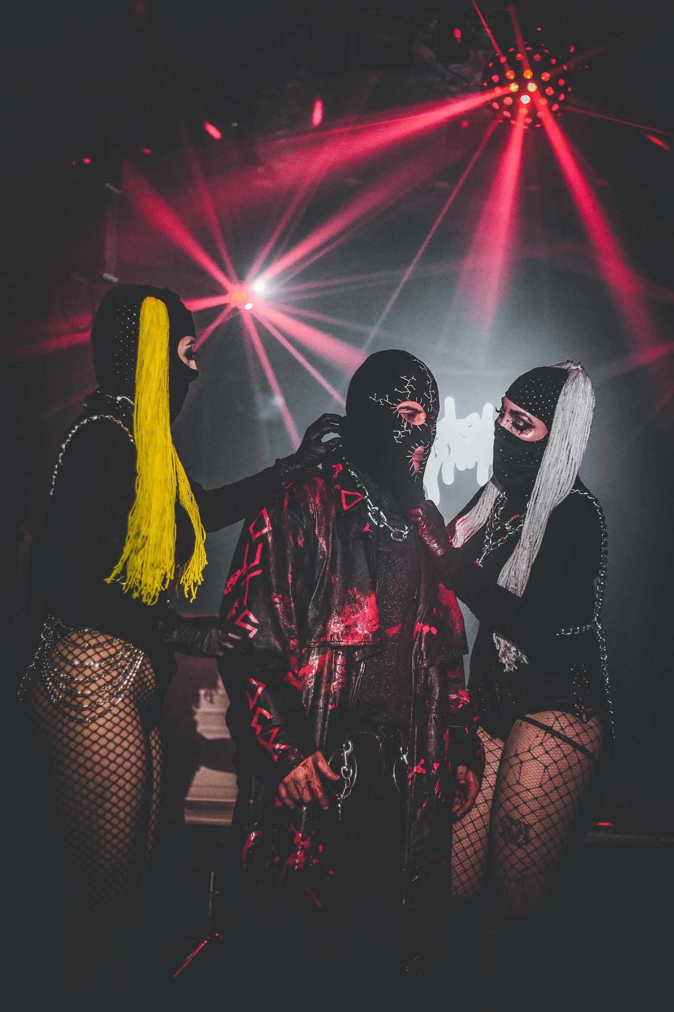 Three people in dark, possibly hooded outfits with red laser lights in the background.