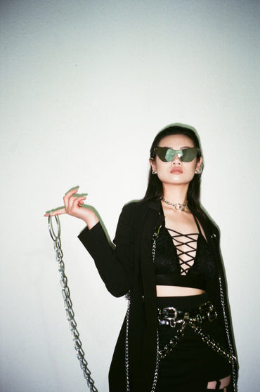 Person wearing a black outfit with sunglasses and holding a chain, against a plain background