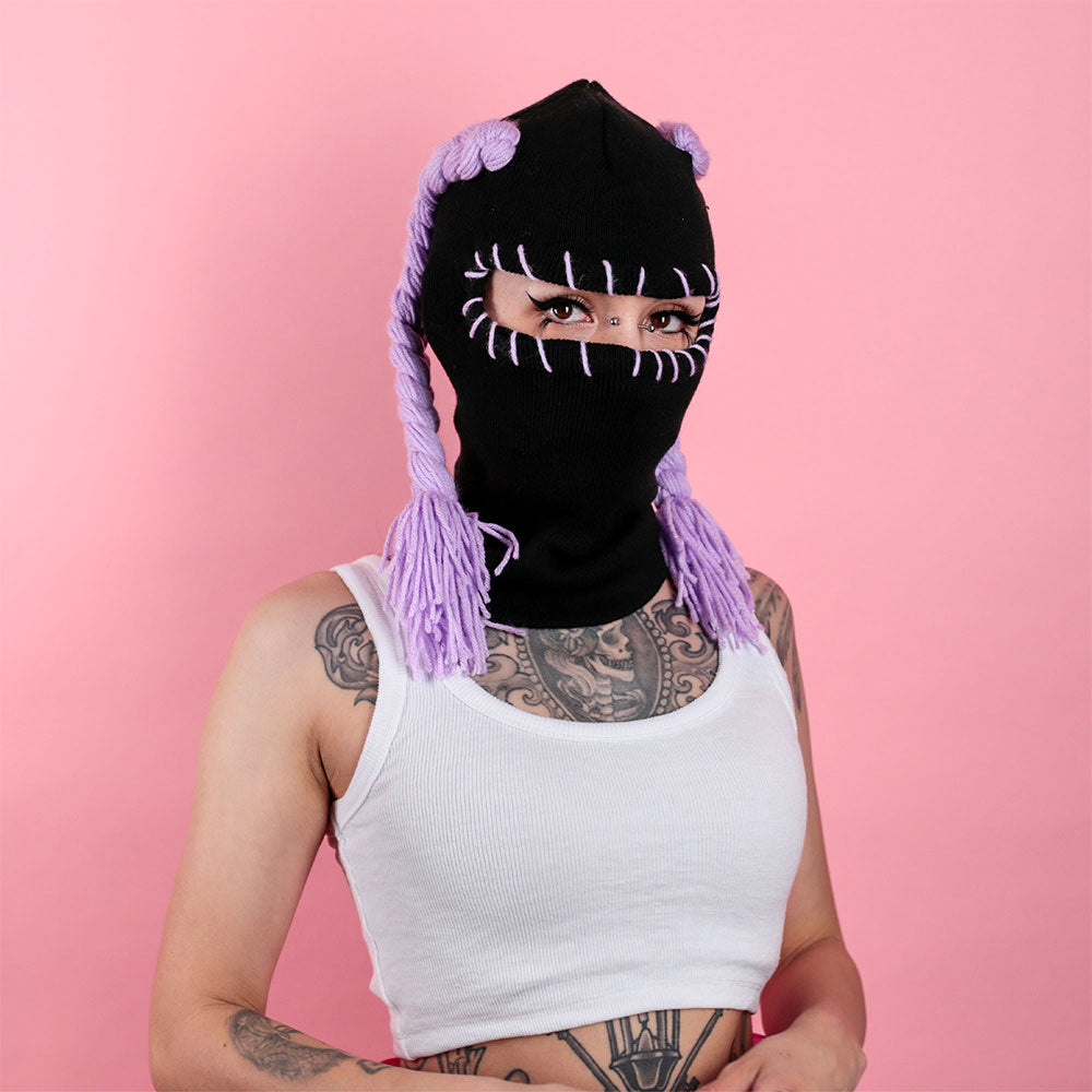 Person wearing a black balaclava with purple braided hair extensions on a pink background