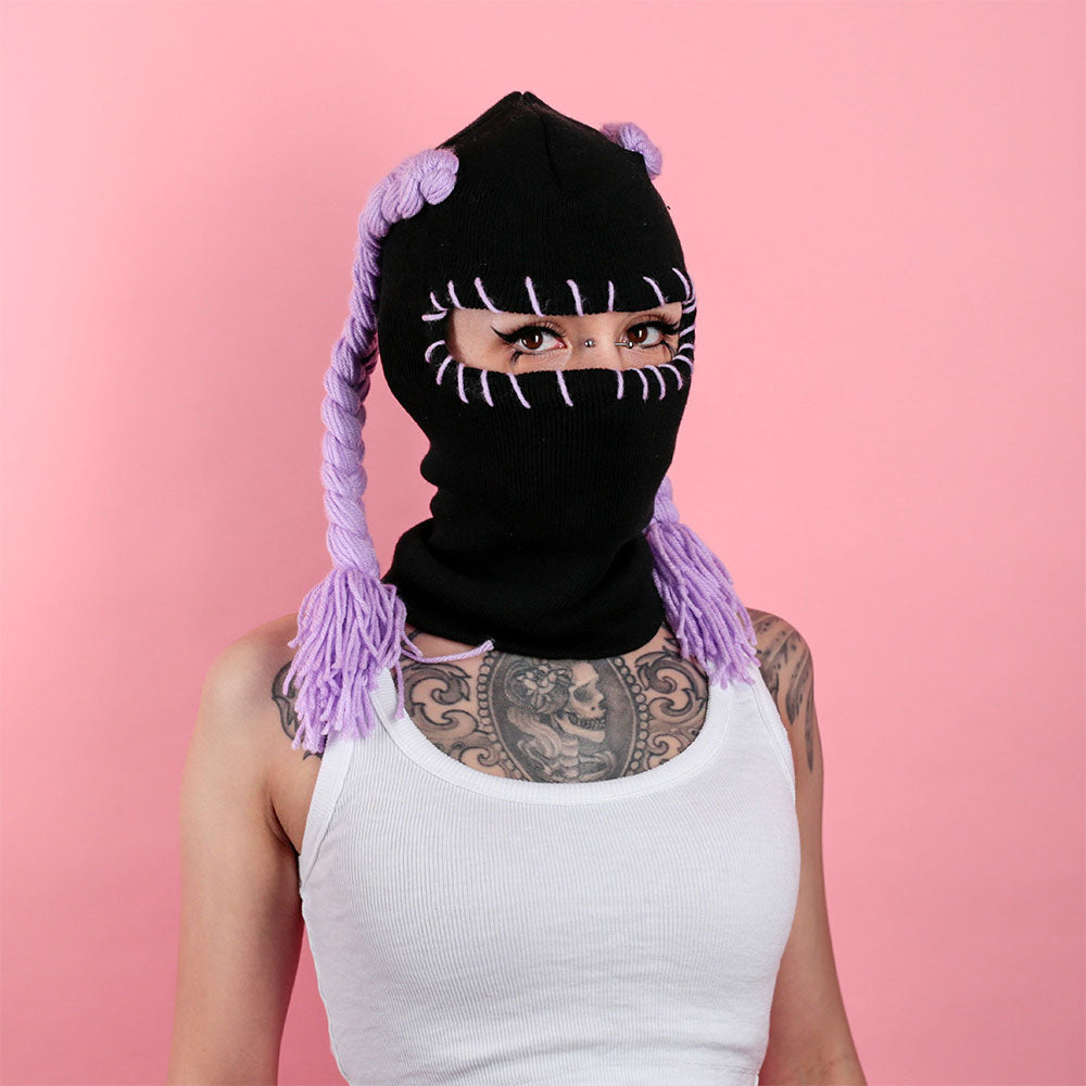 Person wearing a black balaclava with purple pigtails against a pink background