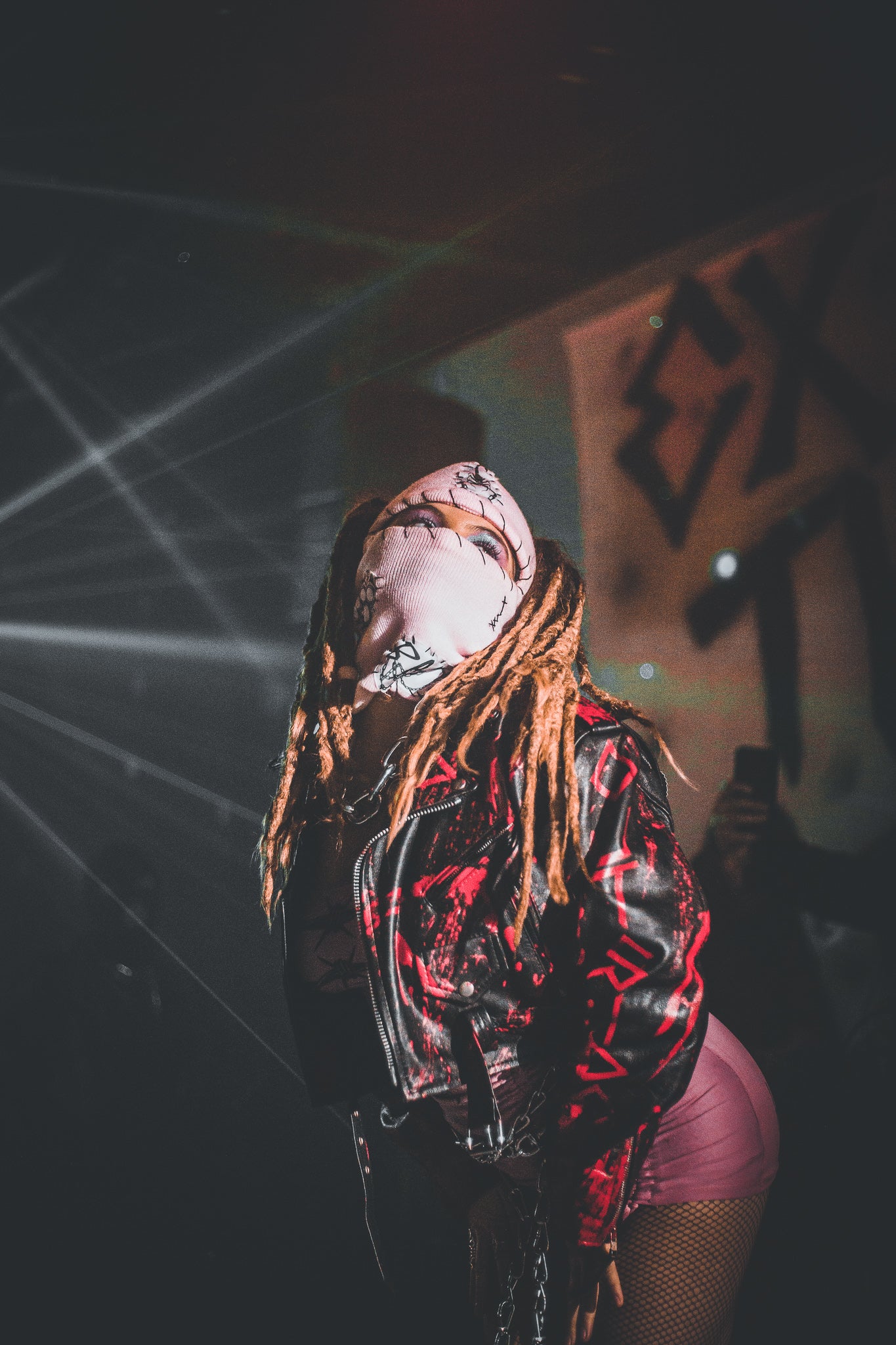 Person with long braided hair wearing a black leather jacket with red patterns on a dark background.