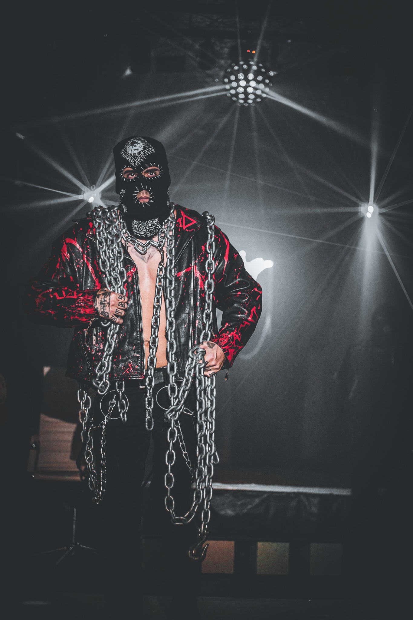 Person wearing a black mask and chains in a dark setting with stage lights.