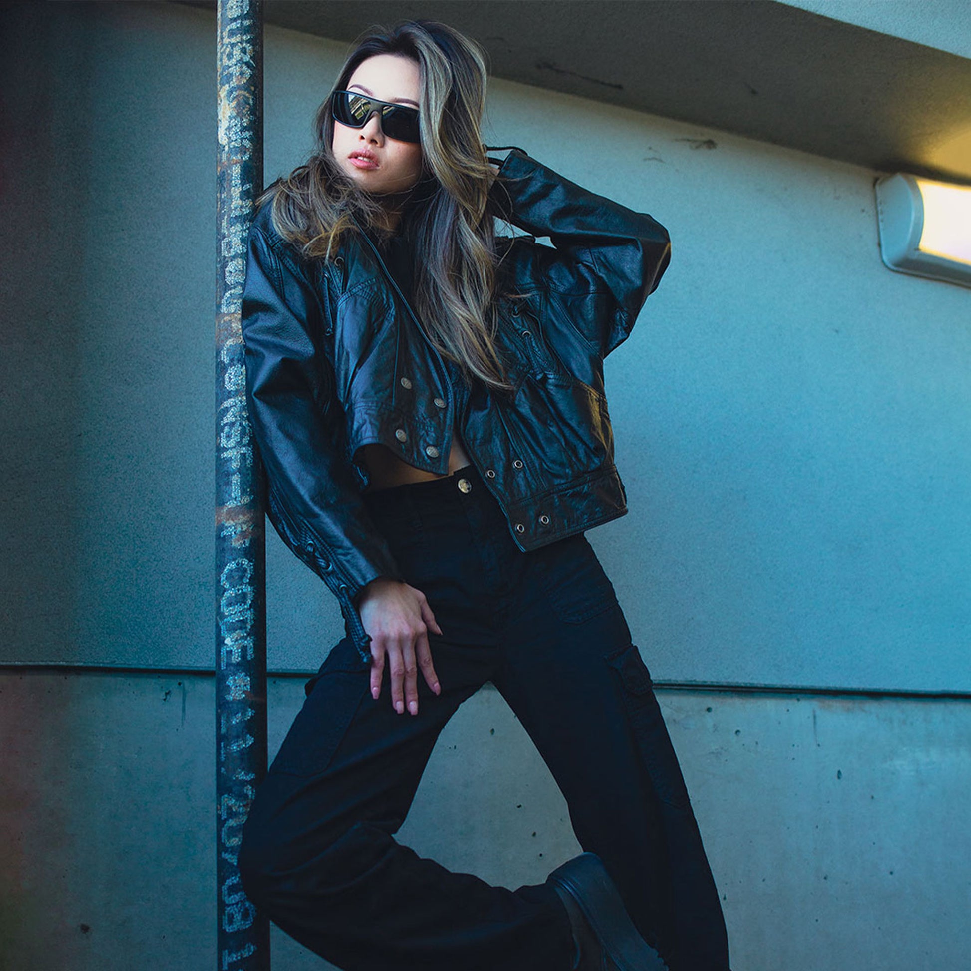 Woman in a green jacket and sunglasses leaning against a wall.