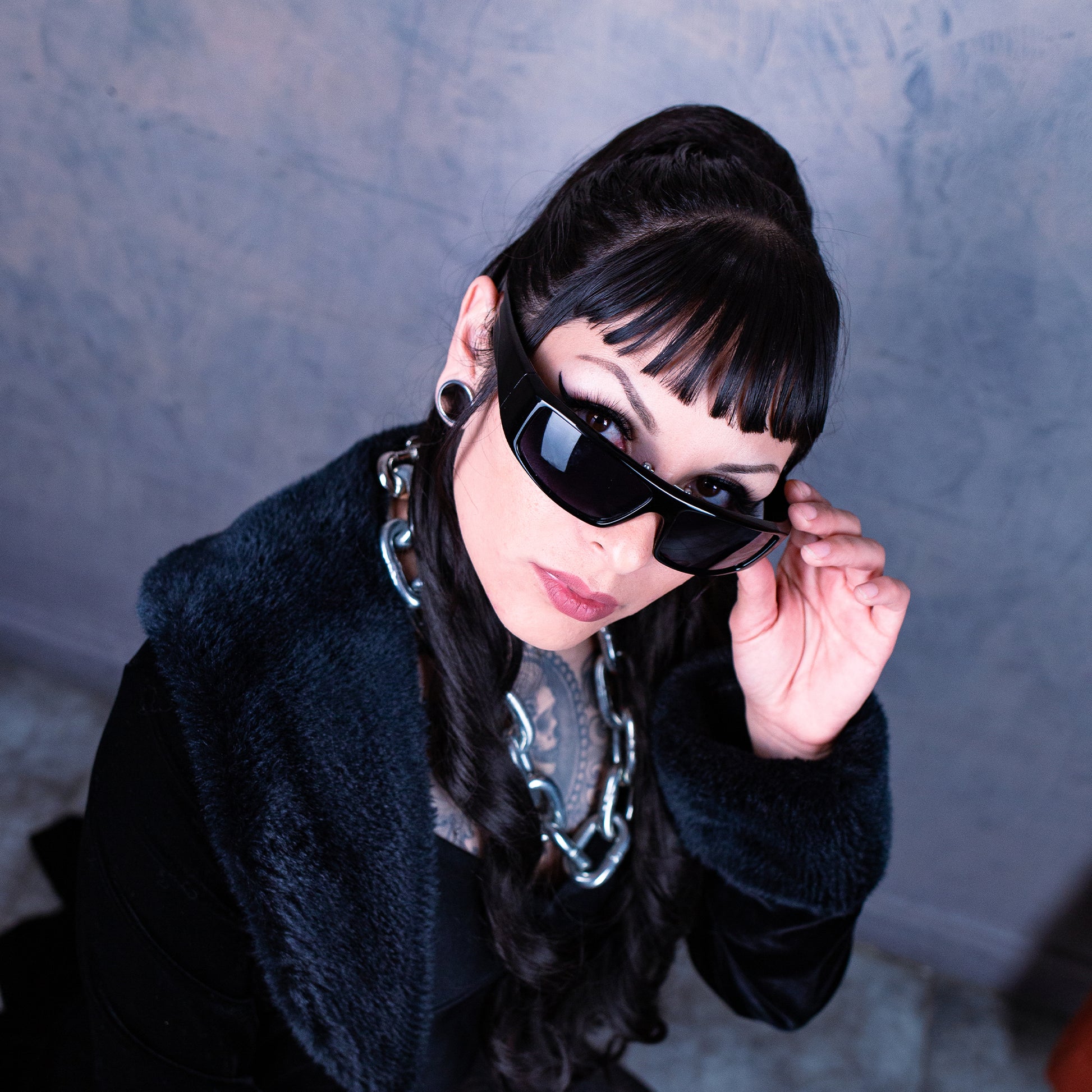 Person wearing sunglasses and a dark coat against a textured gray wall