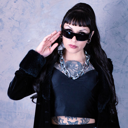 Woman wearing sunglasses and a black outfit with tattoos, standing against a textured gray background