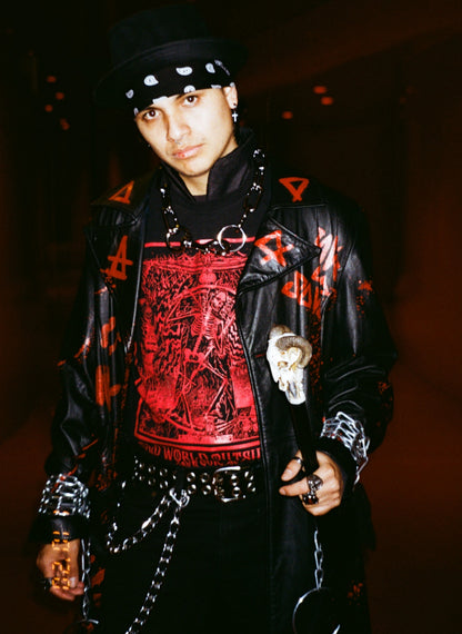 Person wearing a black leather jacket with red designs and a black hat in a dark setting